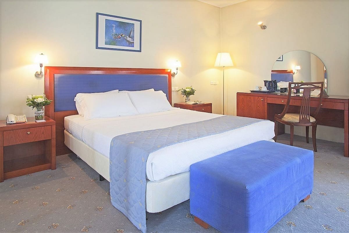 A hotel room with a large bed, bedside tables, a desk with a chair, a desk lamp, a mirror, and a framed picture on the wall.