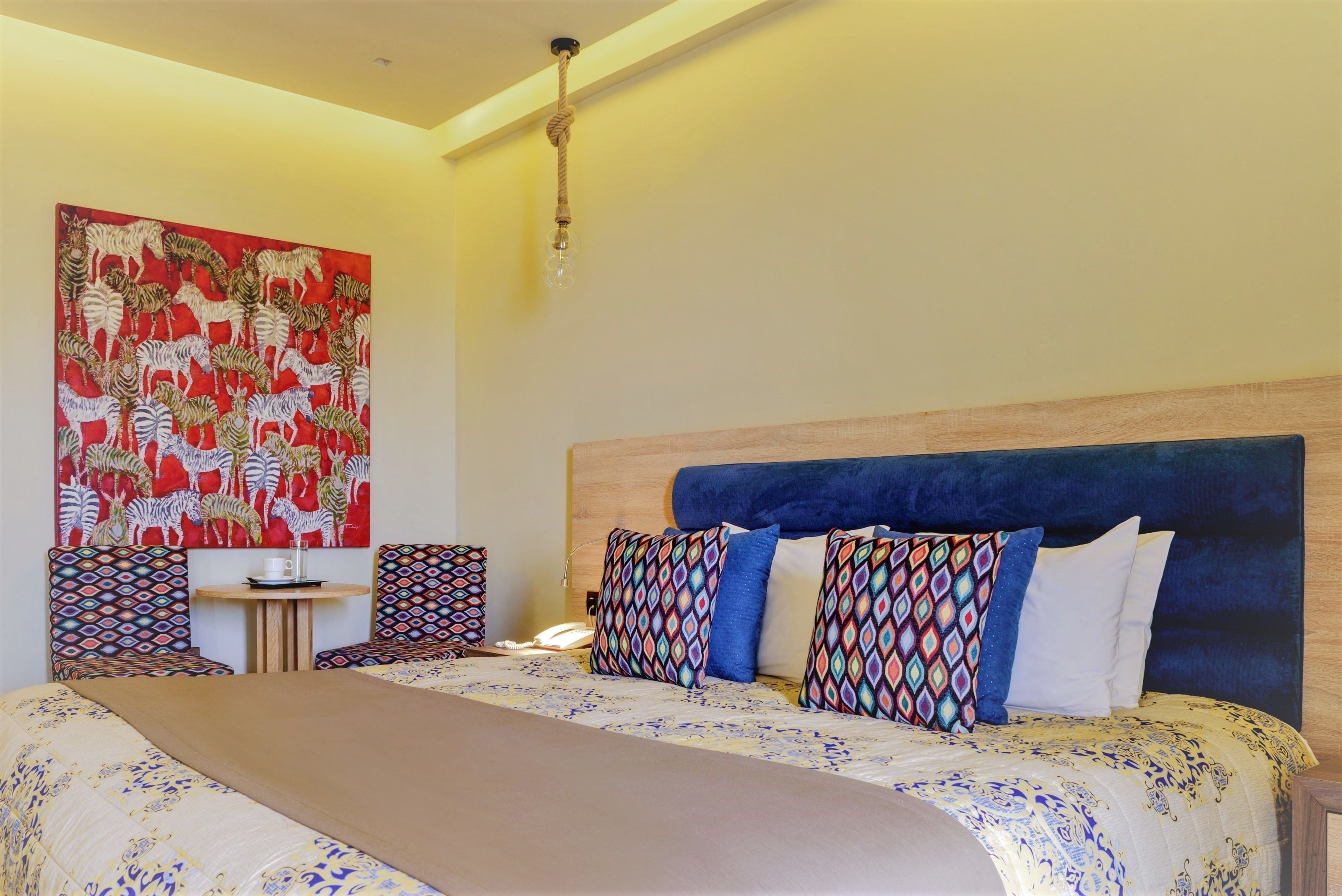 Een hotelkamer met een groot bed, hoofdbord, meerdere kussens, een kleine tafel met een kopje en een kleurrijk abstract schilderij aan de muur.