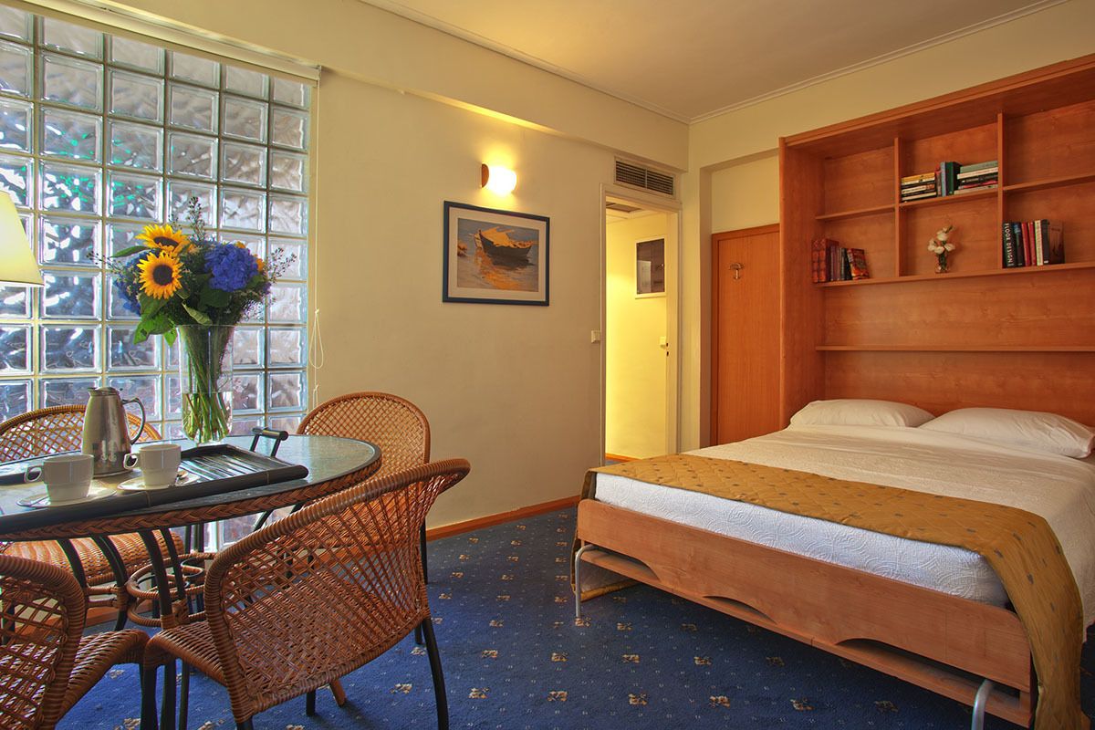 Habitación de hotel con cama, estantería de madera, mesa de comedor con sillas, florero con flores y ventana con vistas.
