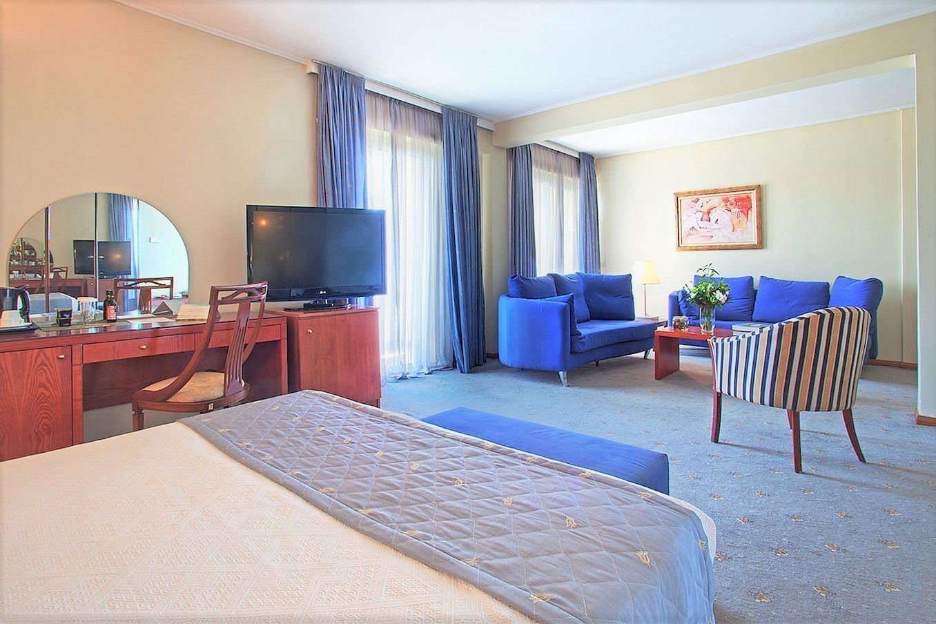 A hotel room with a bed, a desk with a television, a blue sofa, a striped armchair, and a painting on the wall.
