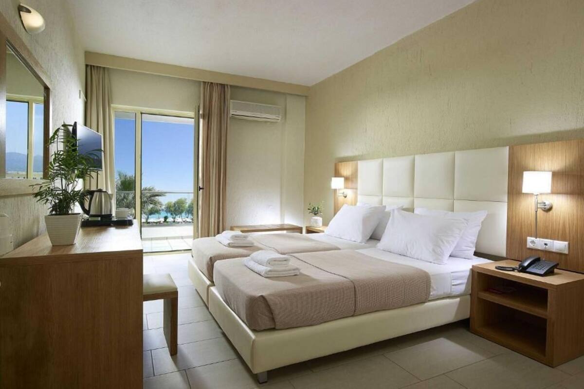 A hotel room with a bed, bedside tables, a desk, a chair, a potted plant, and a view of the outdoors.