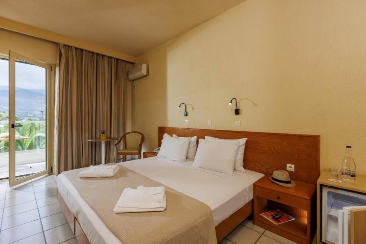 A hotel room with a bed, a nightstand, a chair, a small table, a minibar, and a sliding glass door to the outside.