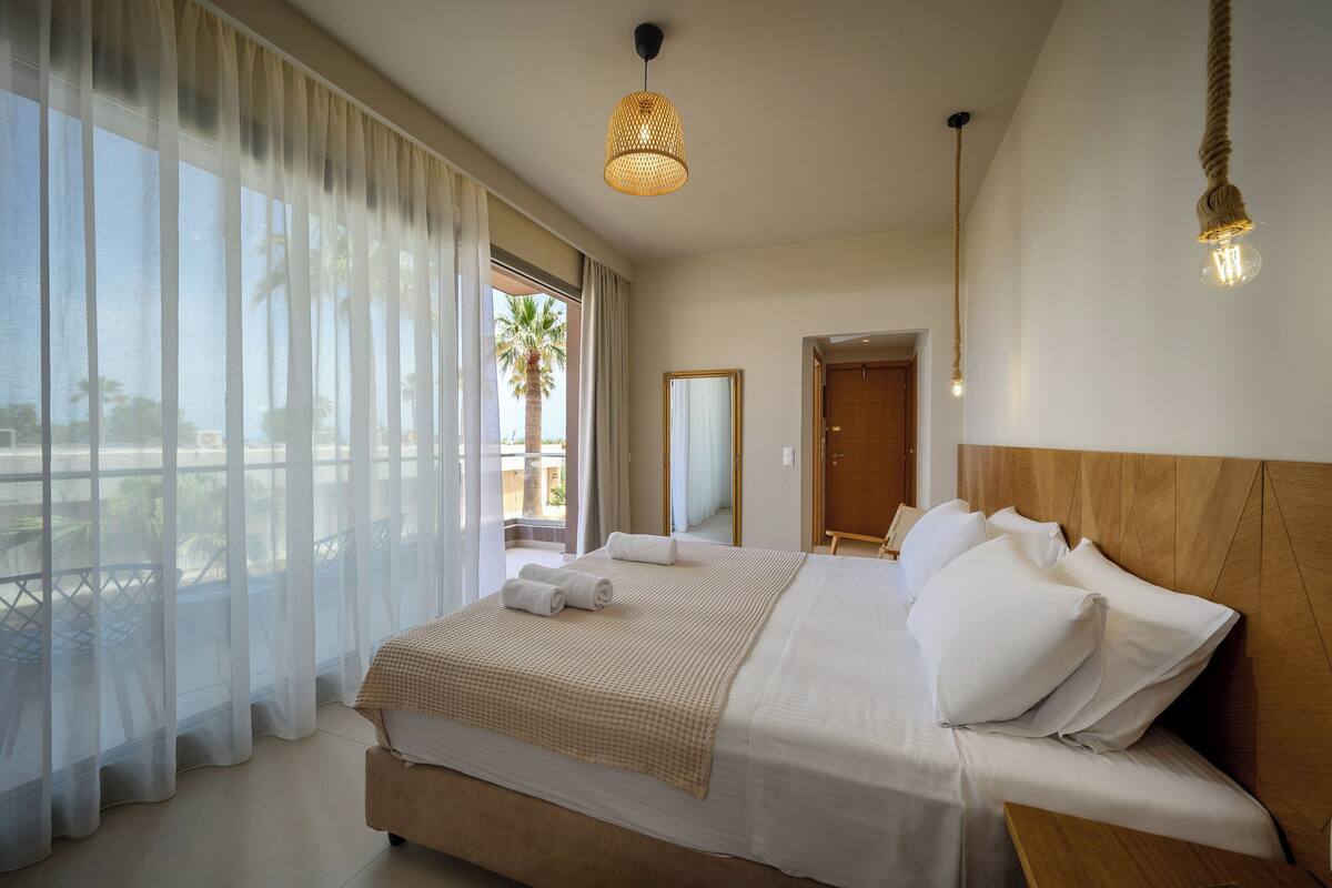 A hotel room with a large bed, a wooden headboard, sheer curtains, a balcony with a view, and a hanging light fixture.
