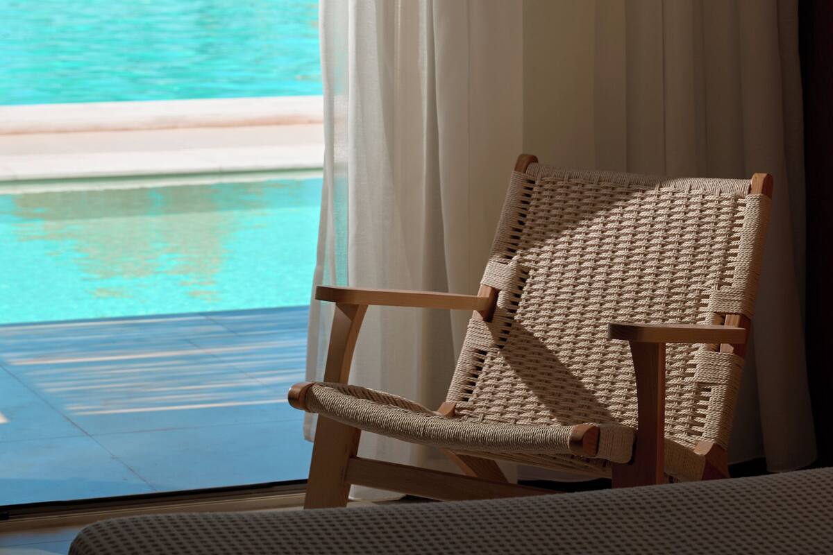 A wicker chair by a window overlooking a pool and beach.