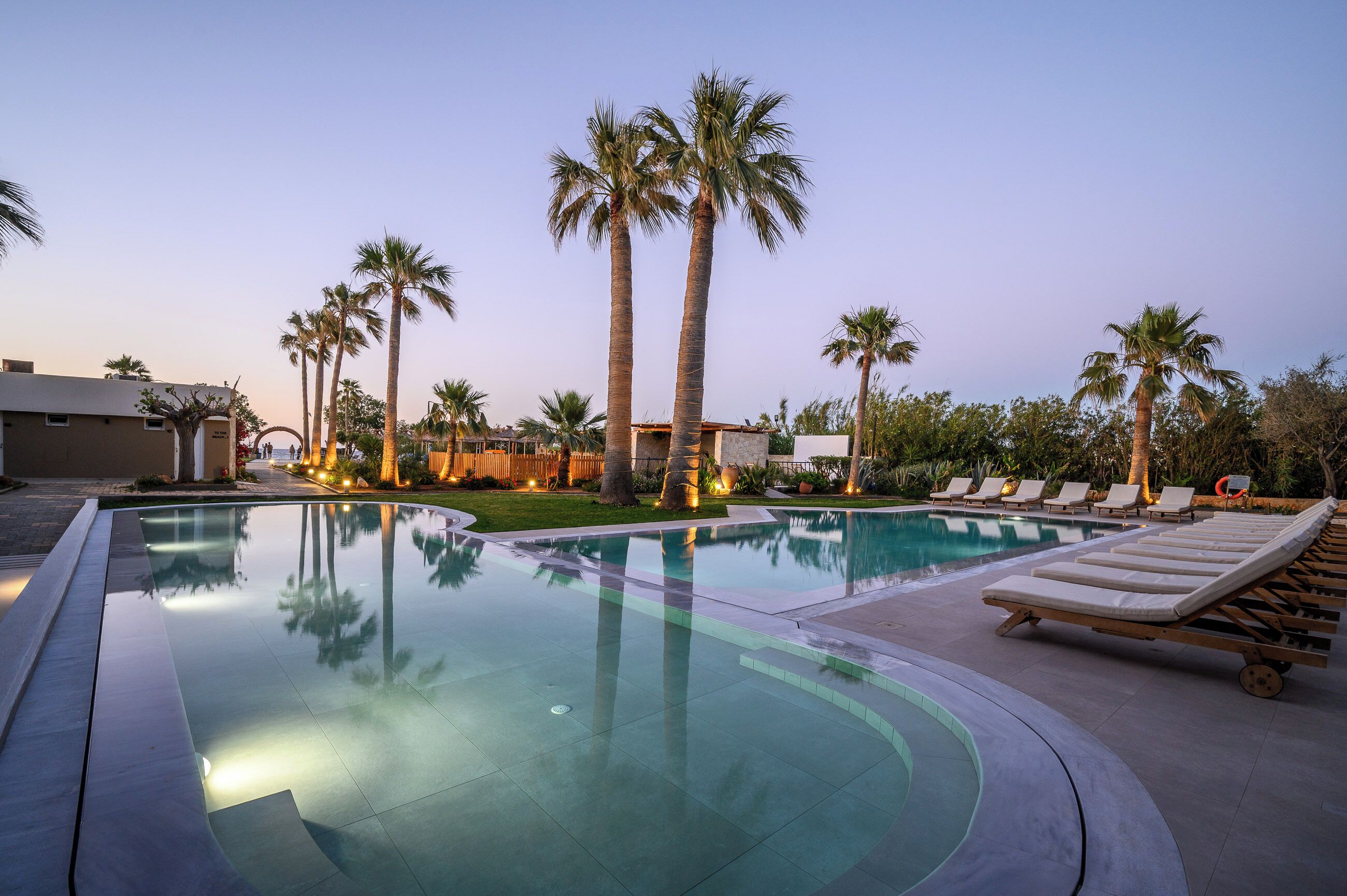 Una piscina con sillones reclinables, palmeras y un edificio al fondo.