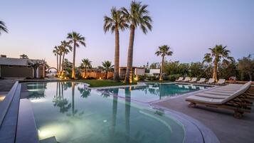 Una piscina con sillones reclinables, palmeras y un edificio al fondo.