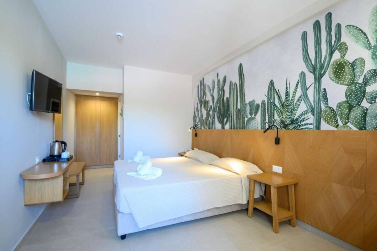 A hotel room with a bed, a wooden headboard, a wall-mounted TV, a bedside table, and a wall art of cacti.