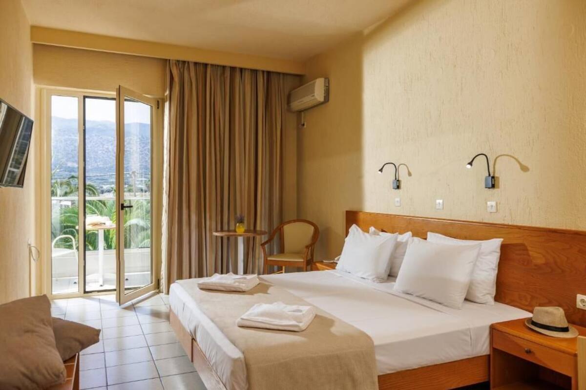A hotel room with a bed, a chair, a small table, a nightstand, and a view through a sliding door.