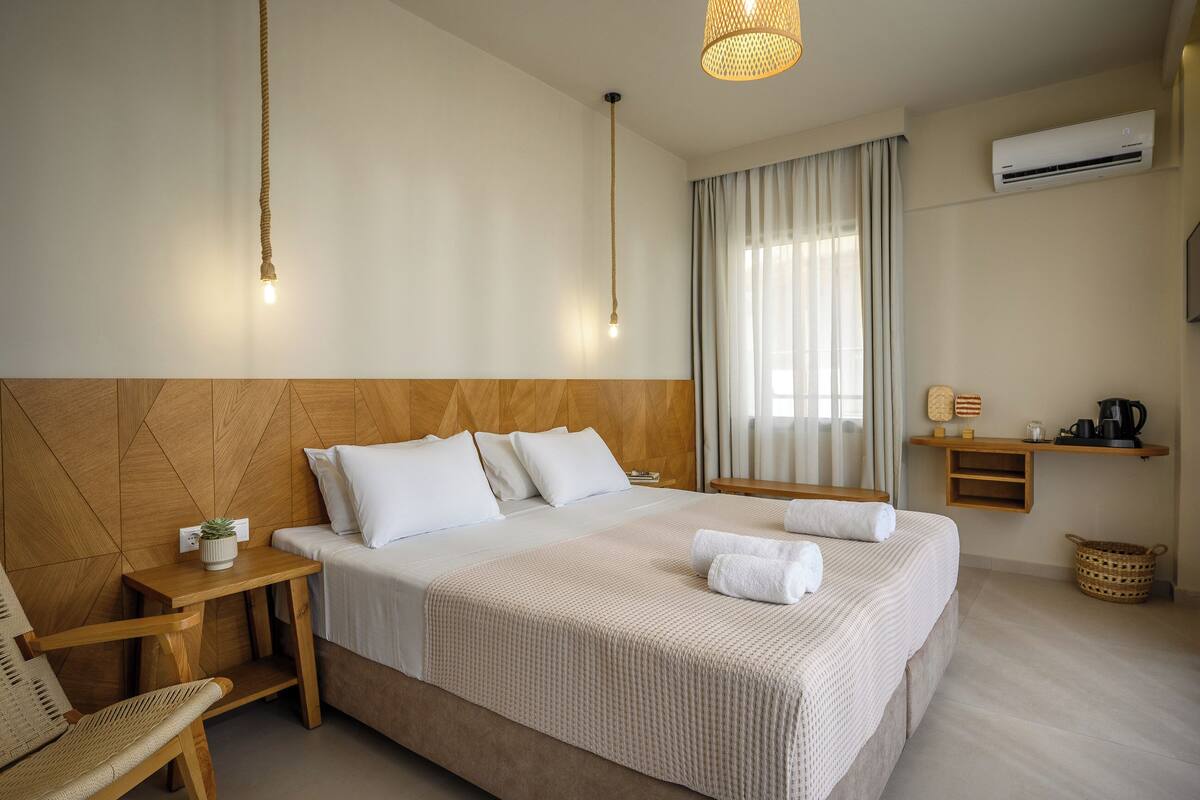 A hotel room with a large bed, bedside tables, a chair, a hanging light fixture, a window with curtains, an air conditioning unit, and a shelf with a kettle.