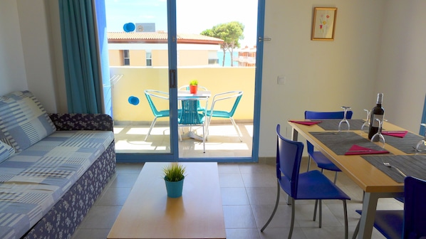 A furnished room with a sofa, a dining table with chairs, a small table with a plant, and a view of the outside balcony area.