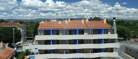 A multi-story building with a red-tiled roof, blue columns, and a swimming pool area with outdoor seating.