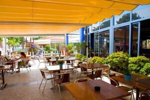 An outdoor café with wooden tables and chairs, potted plants, and a colorful play area in the background.