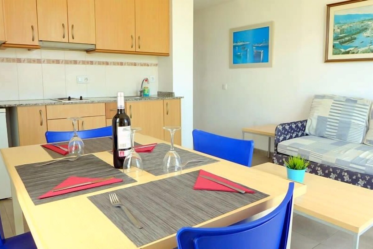 Un comedor con una mesa preparada para dos personas, una botella de vino y copas de vino. Se observan sillas azules y un sofá al fondo.