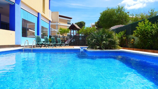 Una piscina con sillas de descanso y un área de estar cubierta, rodeada de vegetación y un edificio de paredes azules y beige.