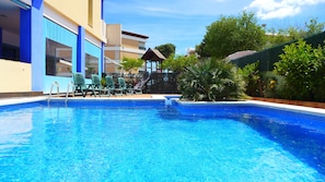 A swimming pool with lounge chairs and a covered seating area, surrounded by greenery and a building with blue and beige walls.