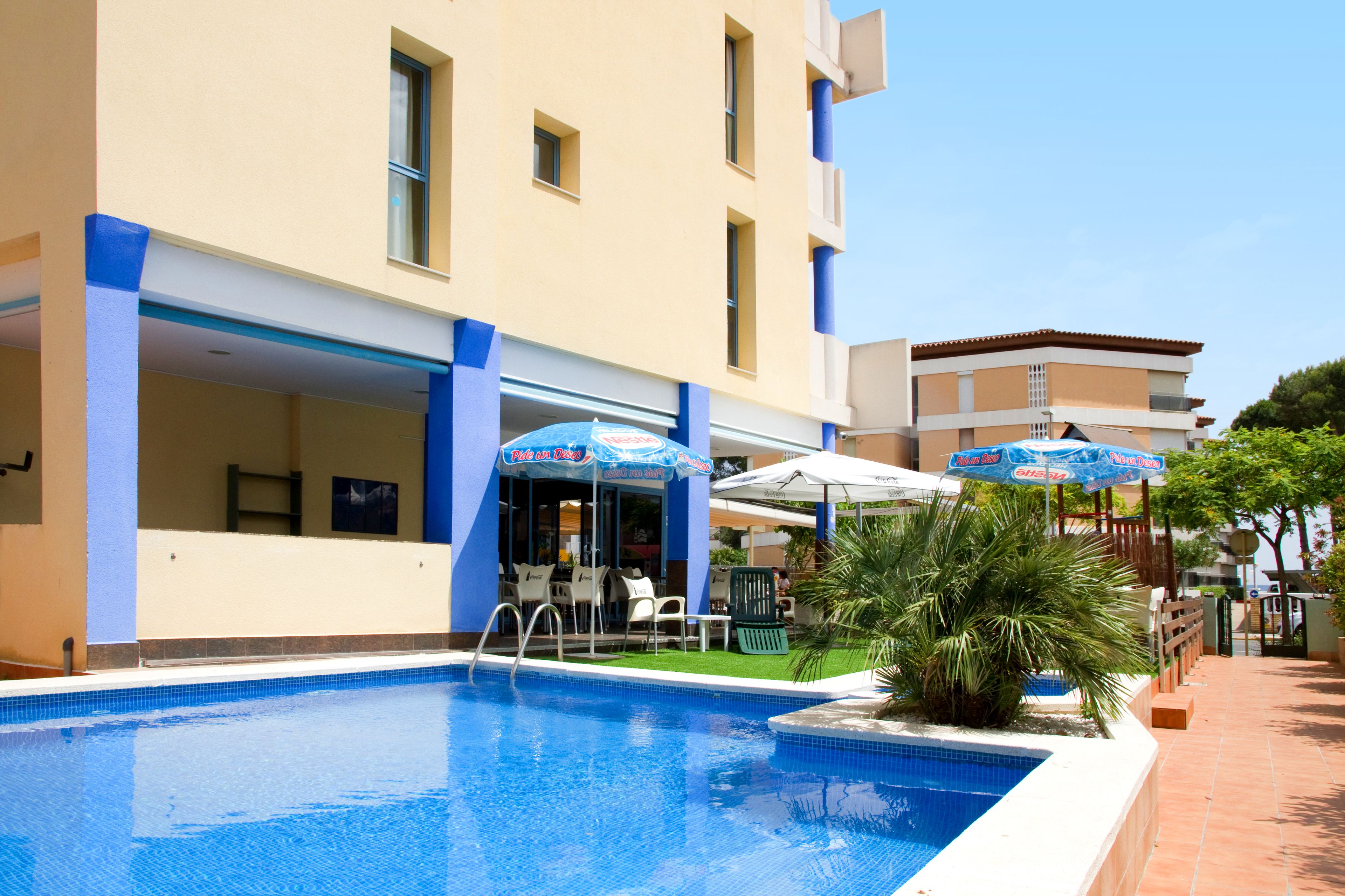 A swimming pool with a blue border, surrounded by a building with blue pillars and a covered seating area with umbrellas.