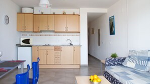 Cocina compacta con armarios de madera, microondas y ventilador de techo. Hay un comedor con mesa y sillas, y un sofá en la sala.