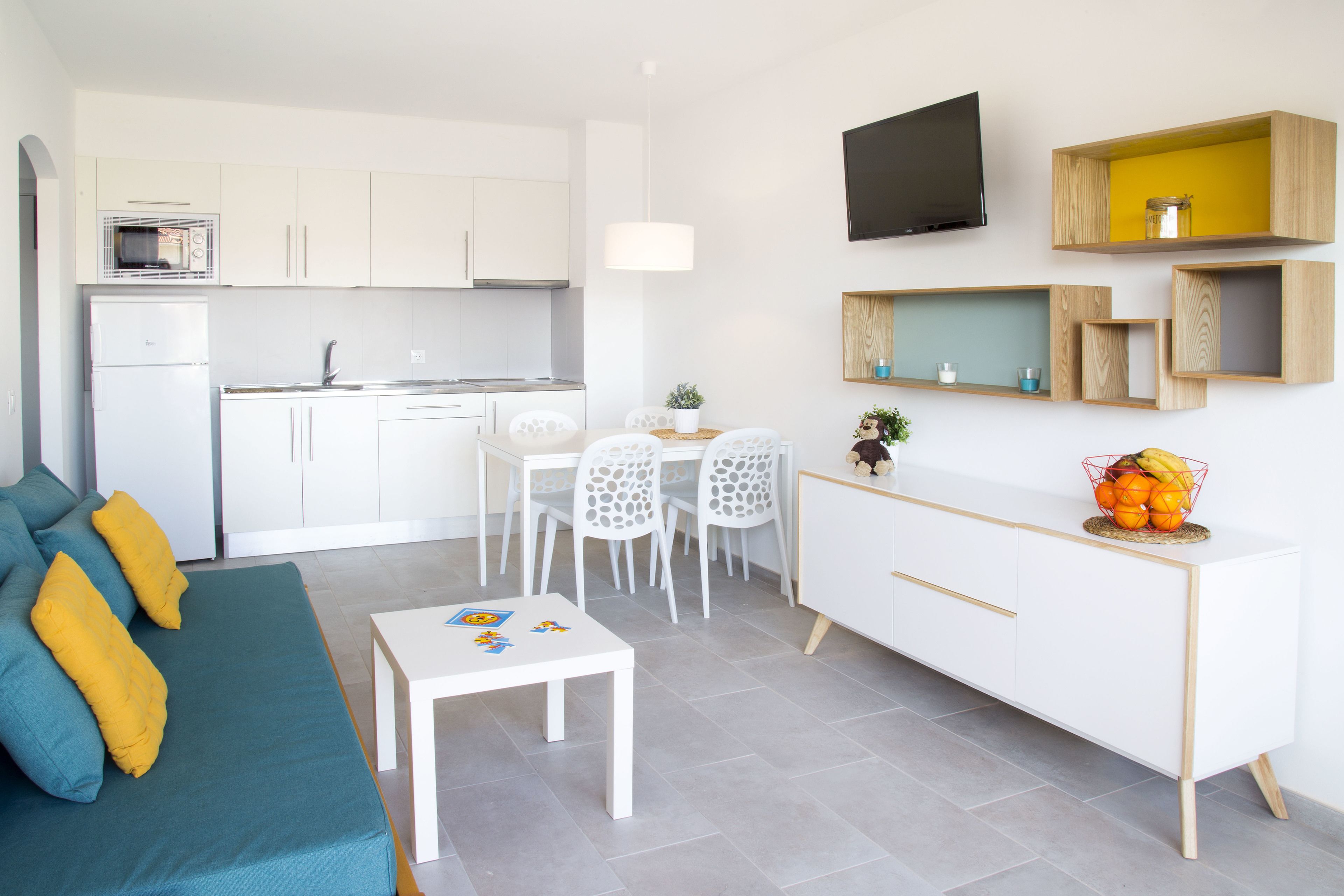 A modern kitchen with white cabinets, a dining area with a white table and chairs, and a living area with a blue sofa and yellow cushions.