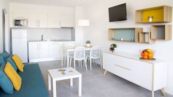 Una cocina moderna con armarios blancos, un comedor con mesa y sillas blancas, y una sala de estar con sofá azul y cojines amarillos.