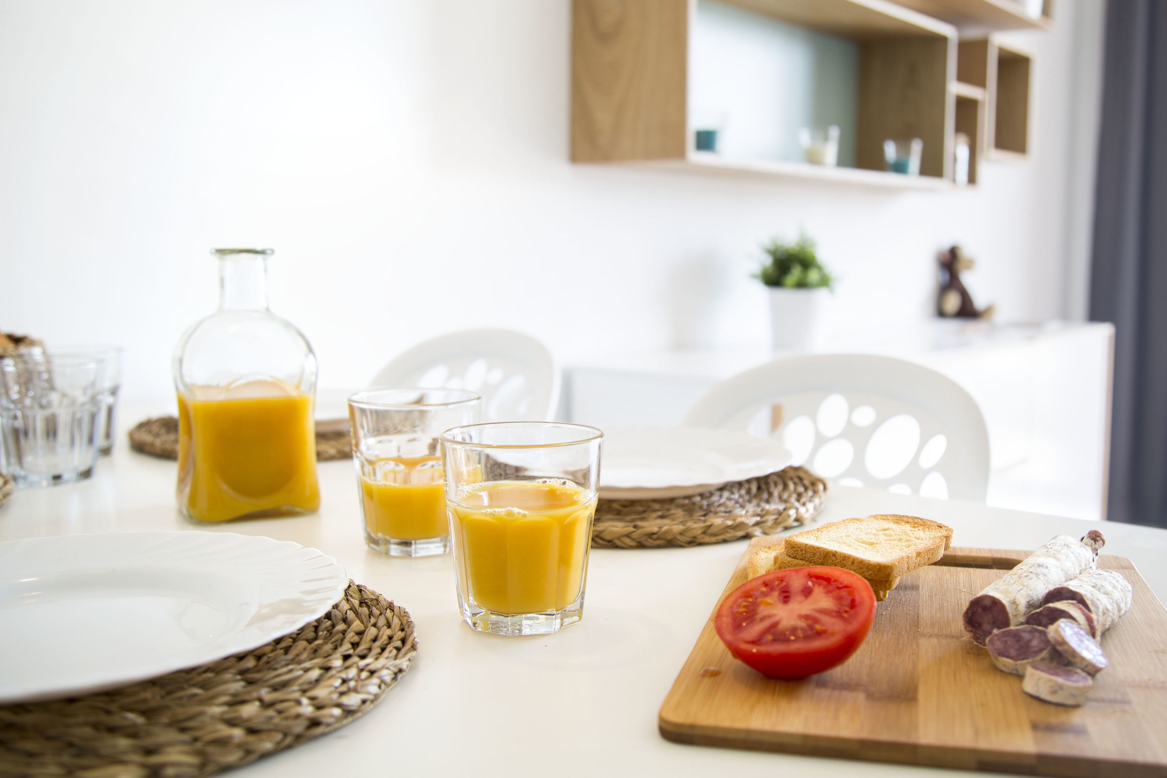 A breakfast setting with a bottle of orange juice, two glasses of orange juice, a plate, a slice of tomato, and a wooden cutting board with sliced sausage.