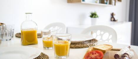 A breakfast setting with a bottle of orange juice, two glasses of orange juice, a plate, a slice of tomato, and a wooden cutting board with sliced sausage.