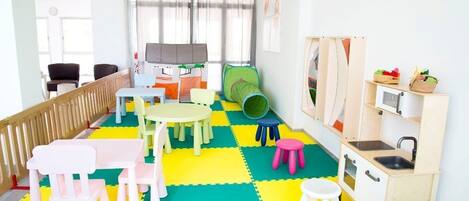 Una sala de juegos infantil con colchonetas coloridas, mesas de juego, sillas y una zona de cocina de juguete.