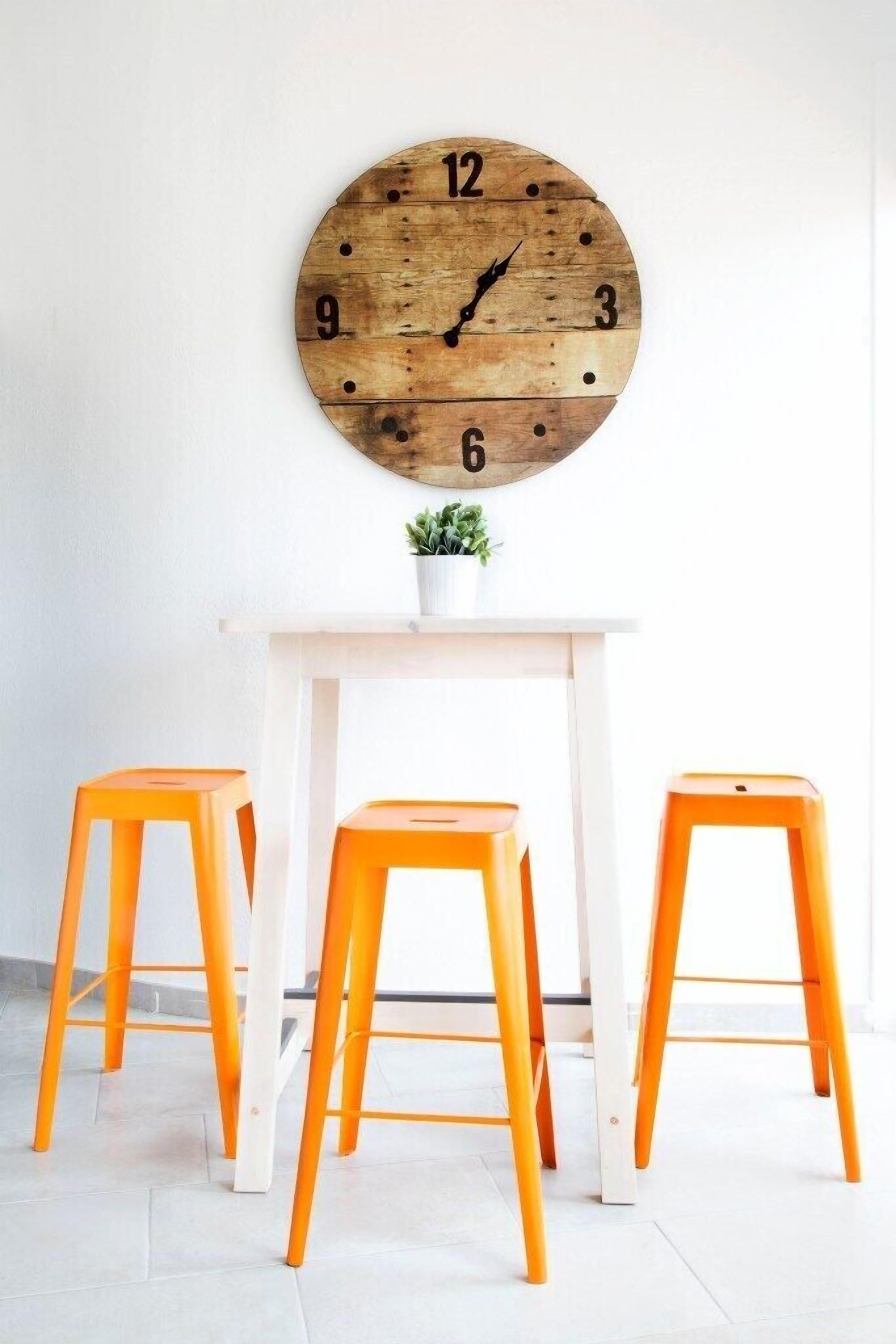 Une horloge murale ronde en bois avec des chiffres, une table blanche et trois tabourets orange.