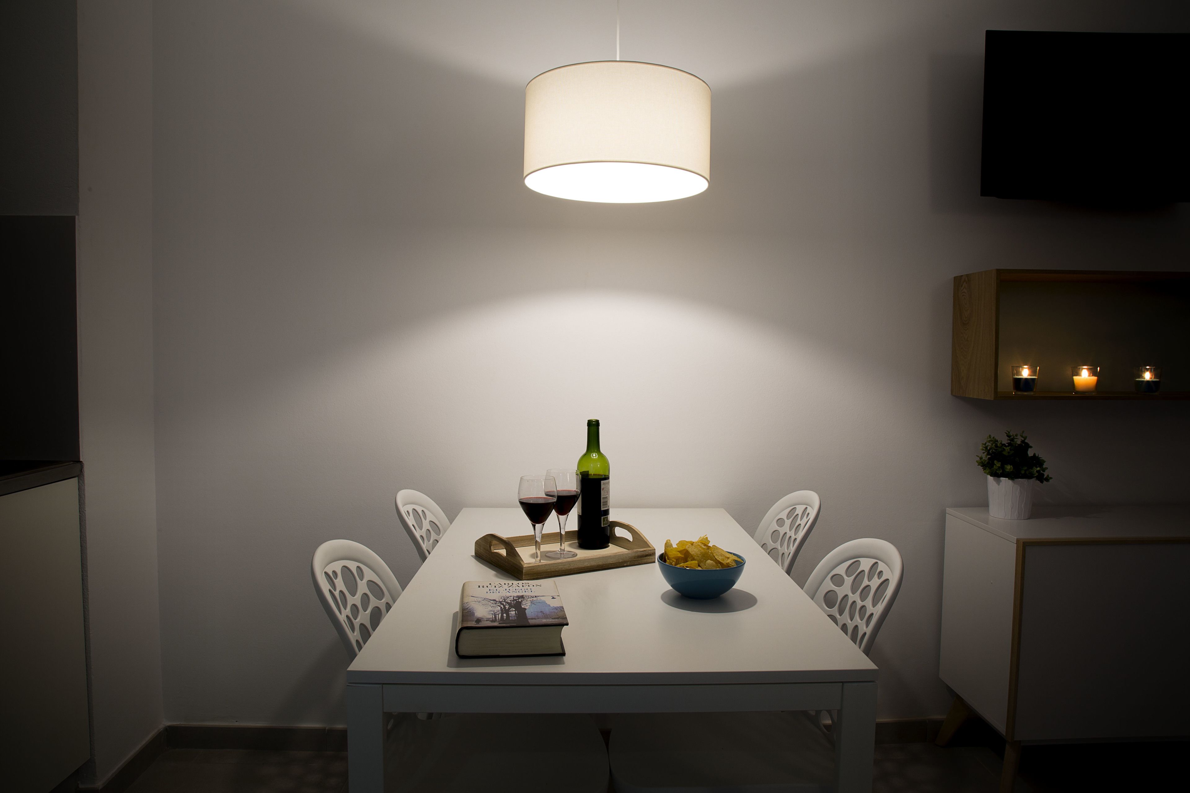 Un comedor con una mesa dispuesta para dos, que incluye una botella de vino, copas, un tazón de papas fritas y un libro.