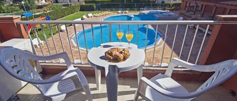 Un balcon avec une piscine, des chaises en plastique blanches et une table garnie de nourriture et de boissons.