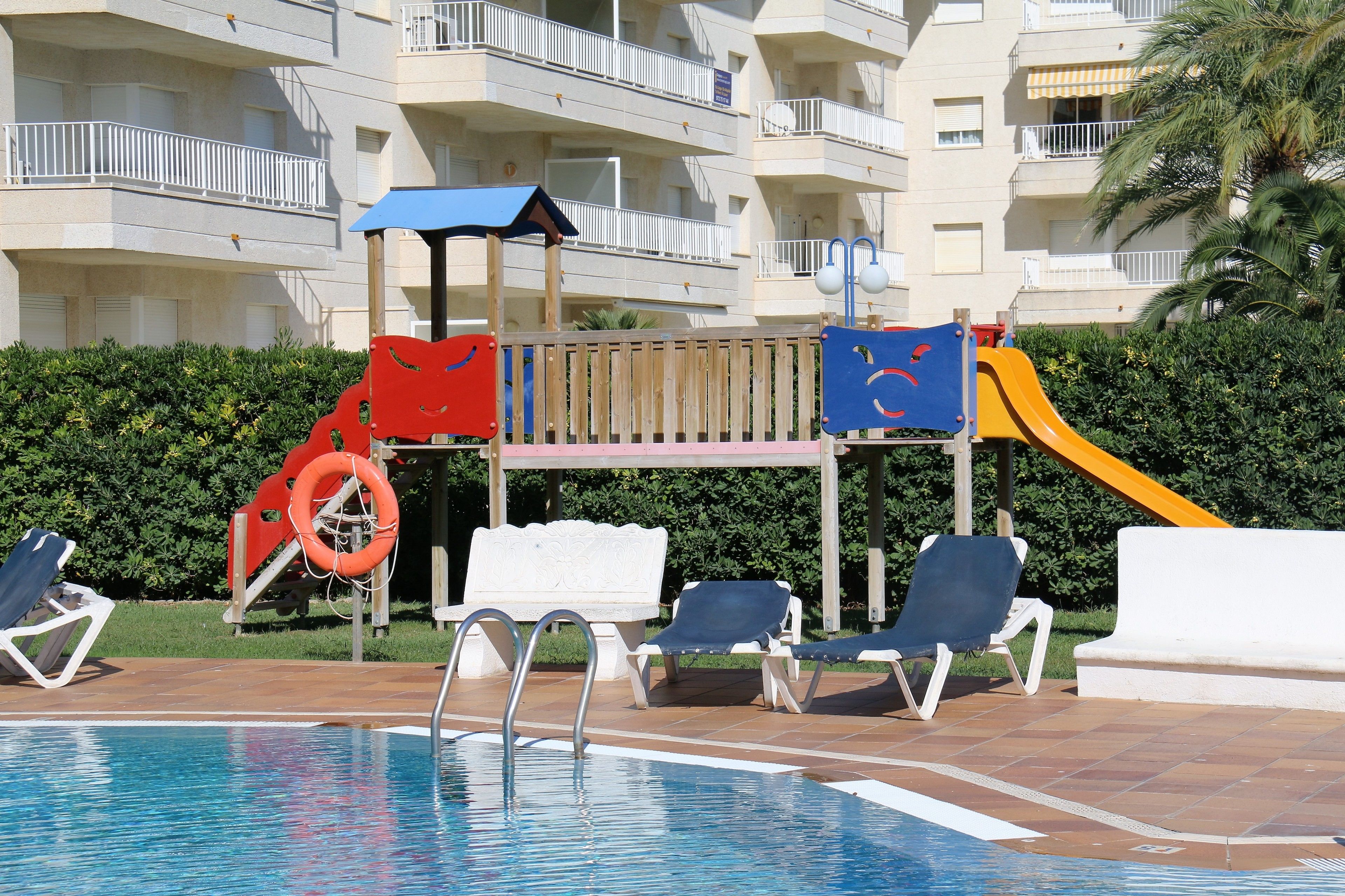 A swimming pool with lounge chairs, a children's play structure, and a building with balconies in the background.