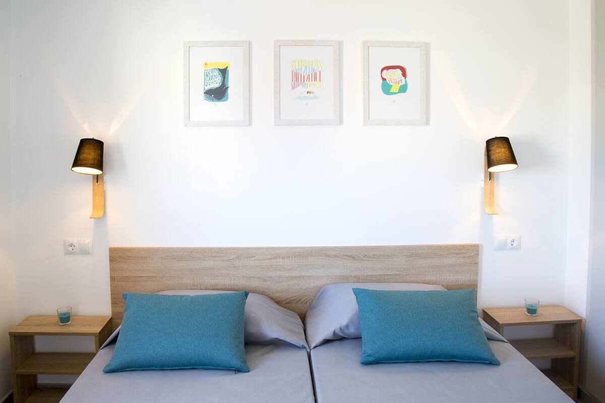 Una cama doble con dos almohadas, un cabecero de madera y dos mesitas de noche con lámparas. Encima de la cama, hay tres obras de arte enmarcadas en la pared.