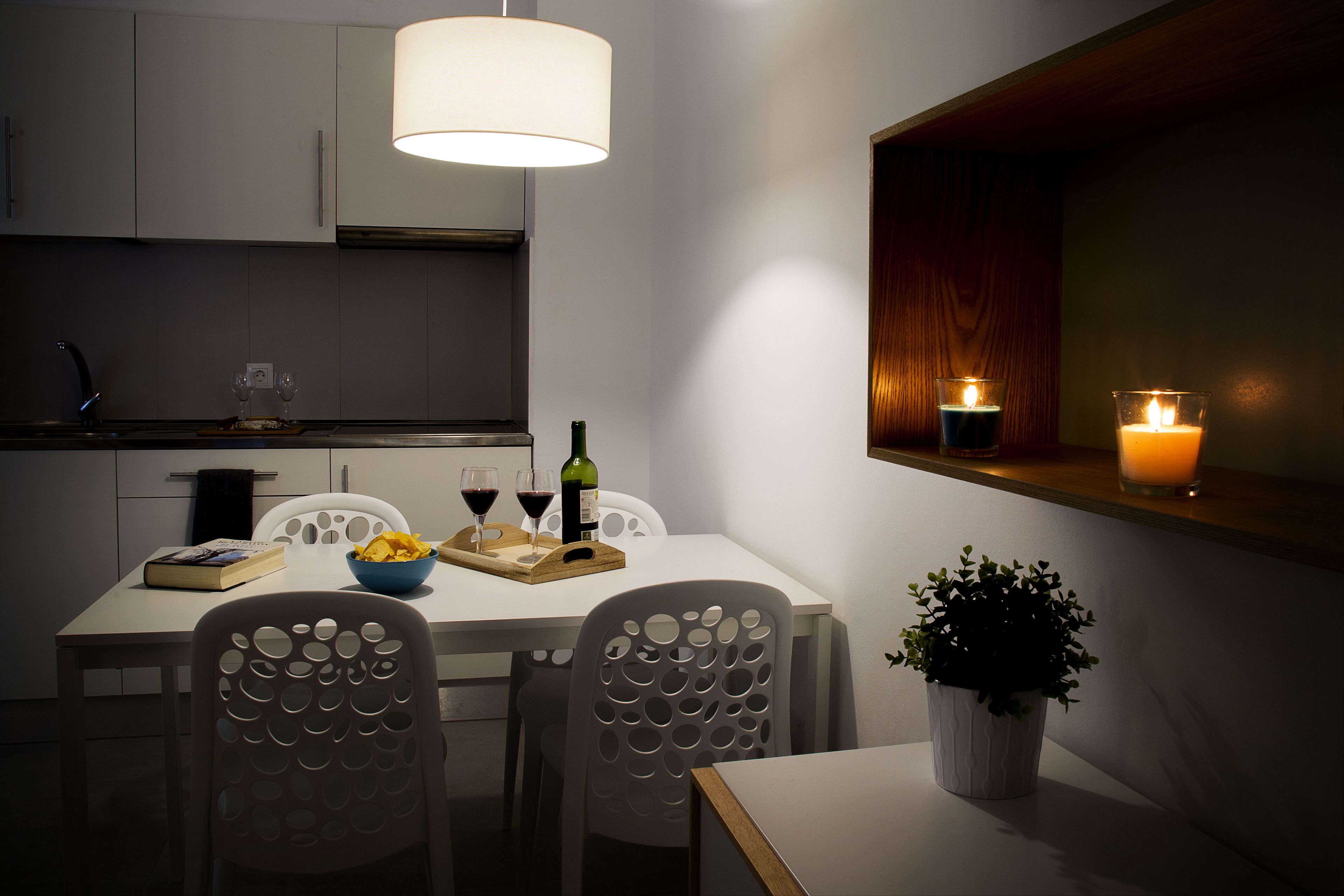 A modern kitchen with white cabinetry, a dining table with chairs, a hanging light fixture, and candles on the windowsill.