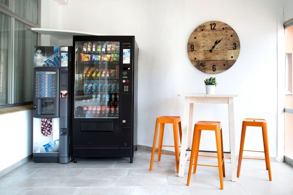 Una máquina expendedora con diferentes bocadillos y bebidas, un reloj de madera en la pared y dos taburetes naranja.