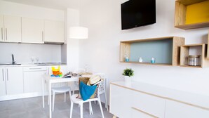 A modern kitchen with white cabinets, a dining table with chairs, a TV mounted on the wall, and wooden shelves above the counter. - RVApartaments Estartit Confort (Torroella de Montgrí)