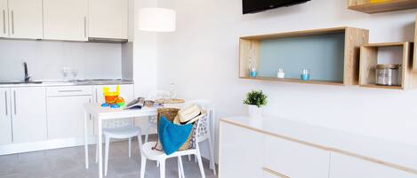 A modern kitchen with white cabinets, a dining table with chairs, a TV mounted on the wall, and wooden shelves above the counter.