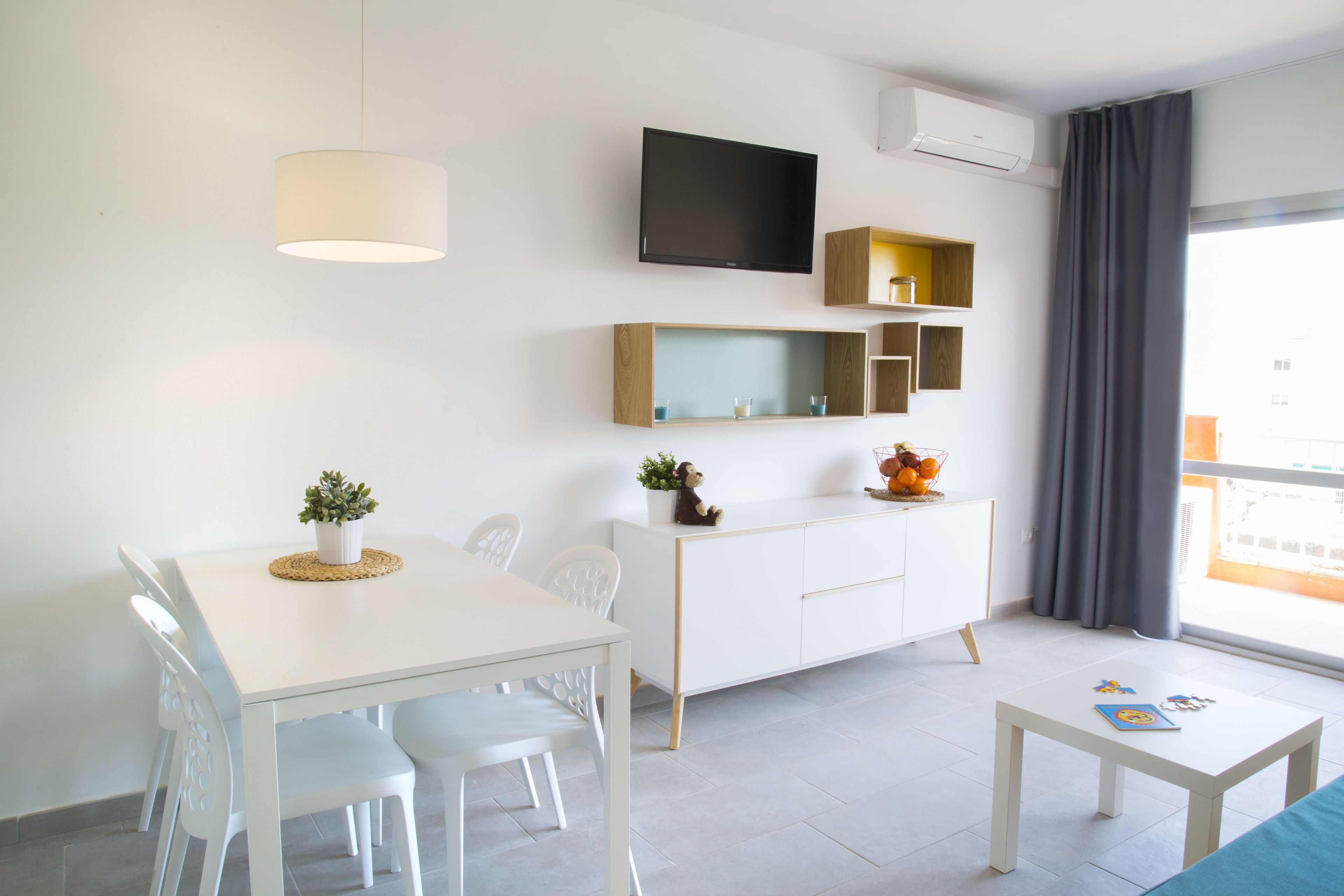 Une pièce moderne et bien éclairée, comprenant une table à manger, des chaises, un canapé blanc, une petite table basse, un téléviseur fixé au mur et une fenêtre avec des rideaux.