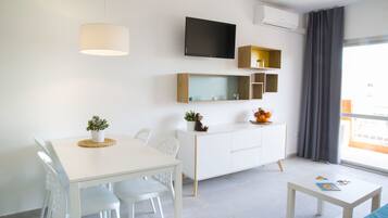 A modern, well-lit room with a dining table, chairs, a white sofa, a small coffee table, a TV mounted on the wall, and a window with curtains.