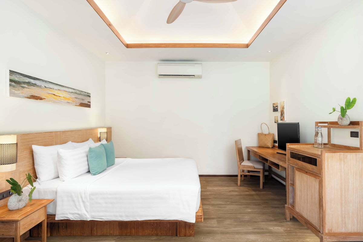Une chambre d’hôtel moderne avec un lit en bois, un bureau et une chaise. On y trouve un ventilateur de plafond, un appareil de climatisation et un tableau au mur.