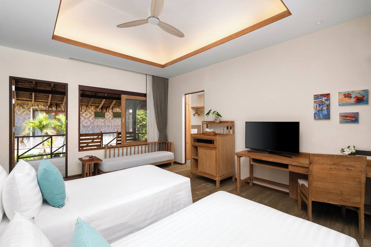 Une chambre d’hôtel avec deux lits simples, un bureau en bois, une télévision et un balcon donnant sur des palmiers.