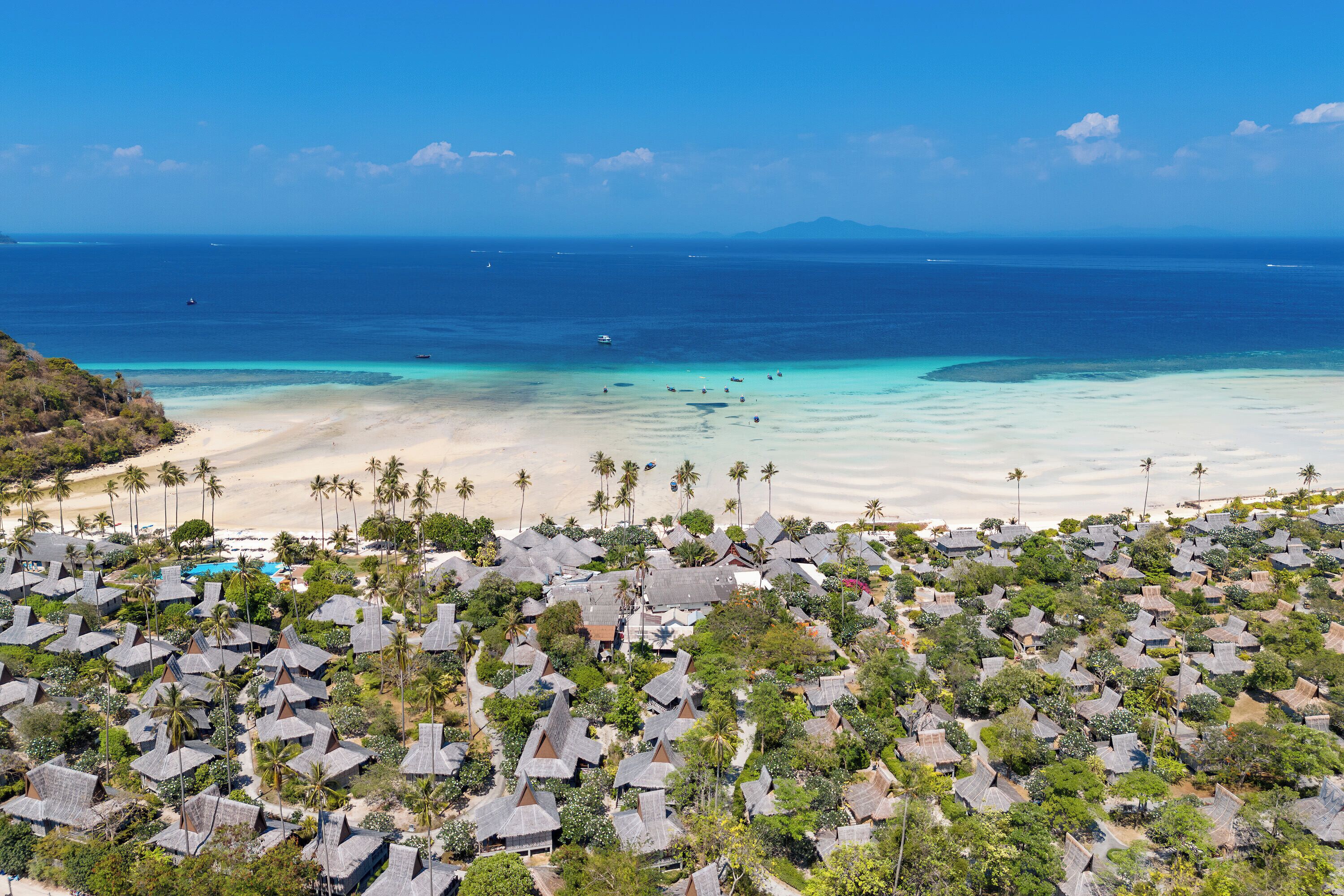 An aerial view of a coastal resort with numerous thatched-roof huts, a sandy beach, and a clear blue sea.