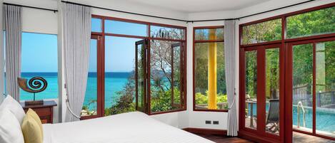 A bedroom with a large bed, a view of the ocean, and a sliding glass door leading to a balcony with a pool.