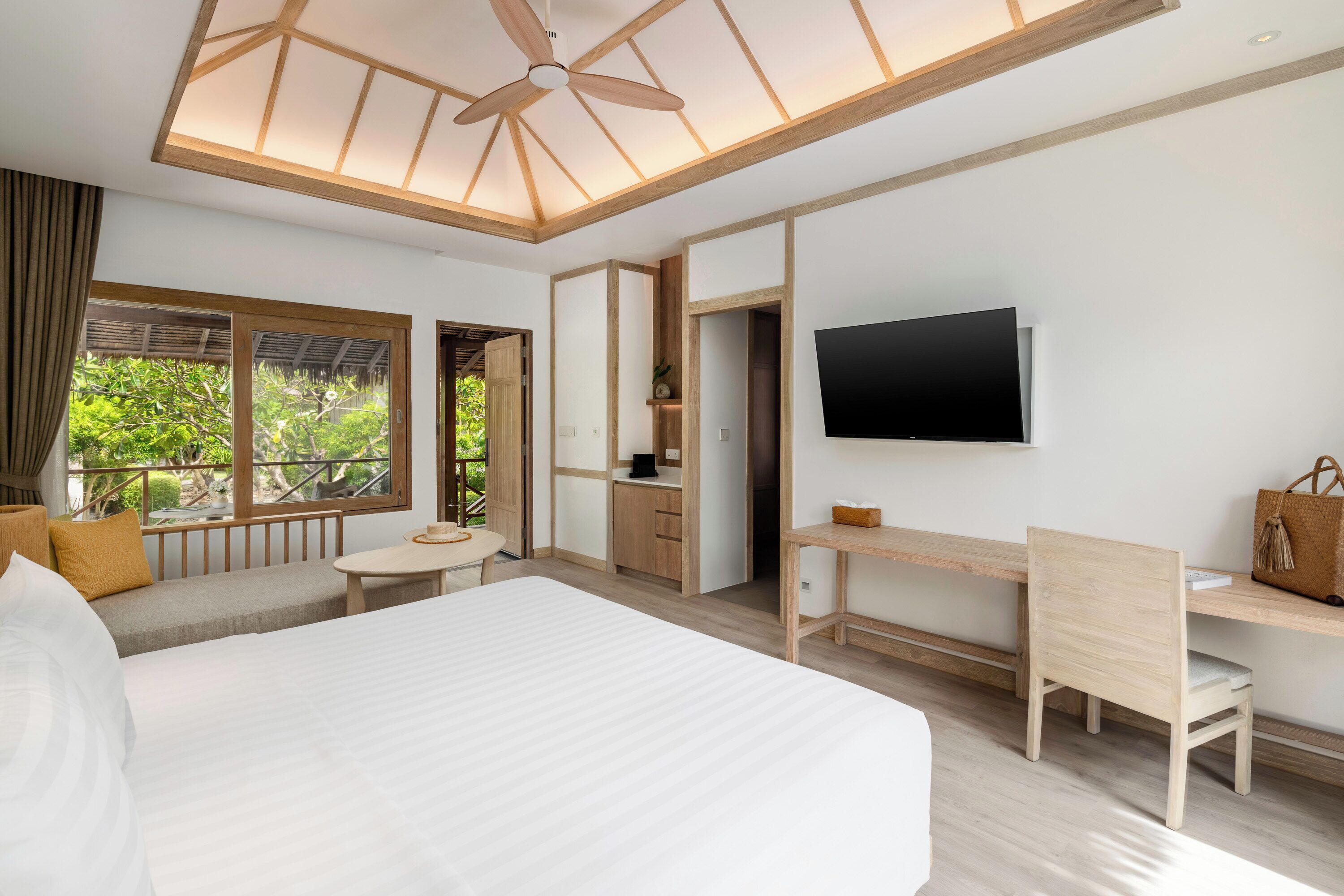 A modern hotel room with a large bed, a desk, a chair, a TV, and a balcony with a view of greenery.