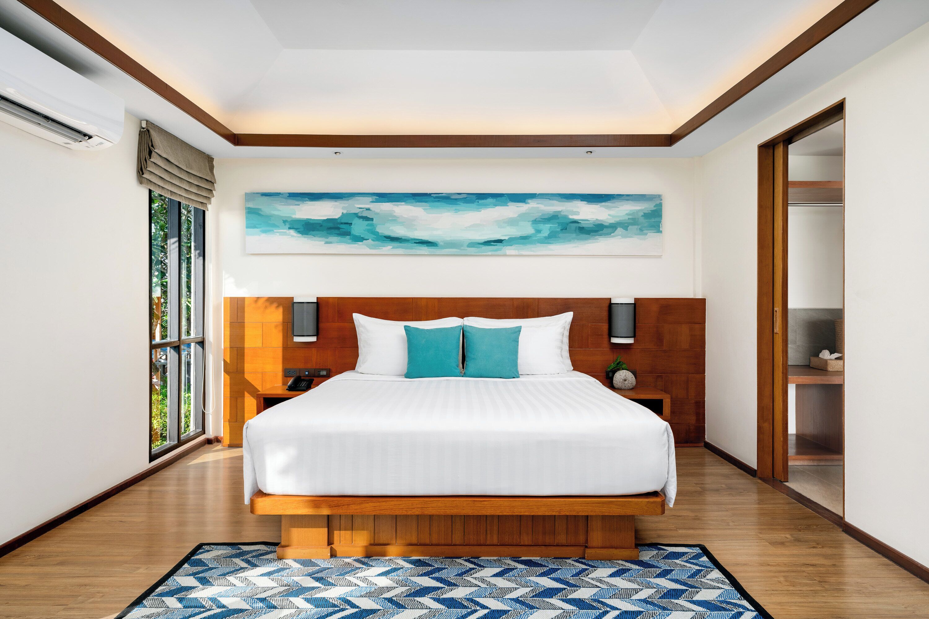 A modern hotel room with a large bed, wooden headboard, a painting above the bed, a nightstand, and a view through a window.