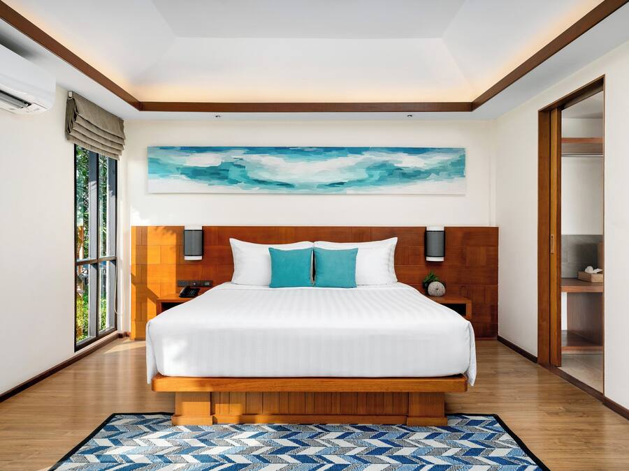 A modern hotel room with a large bed, wooden headboard, a painting above the bed, a nightstand, and a view through a window.