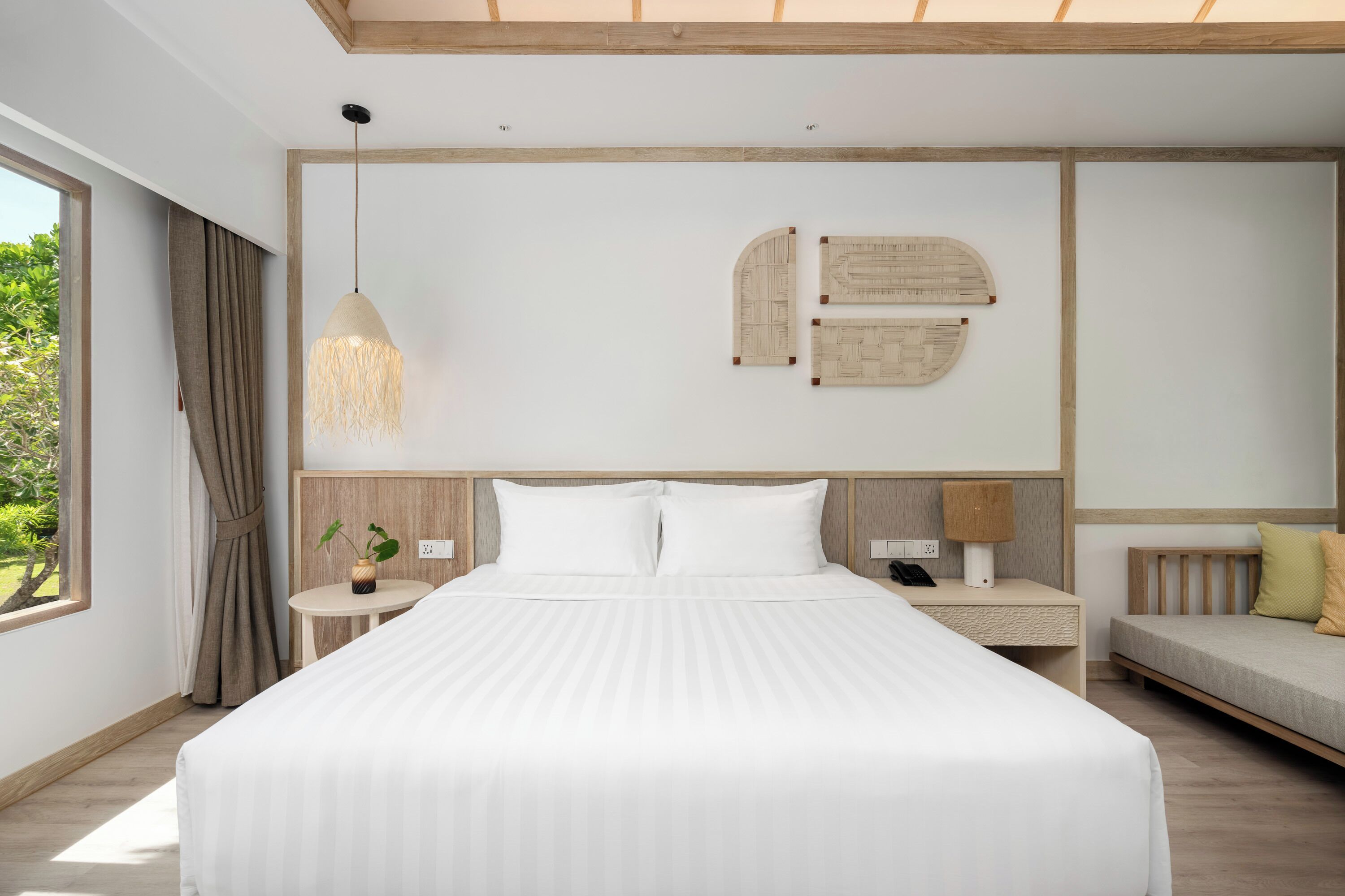 A modern hotel room with a large bed, a bedside table with a lamp, a small side table with a plant, and a sofa with cushions.