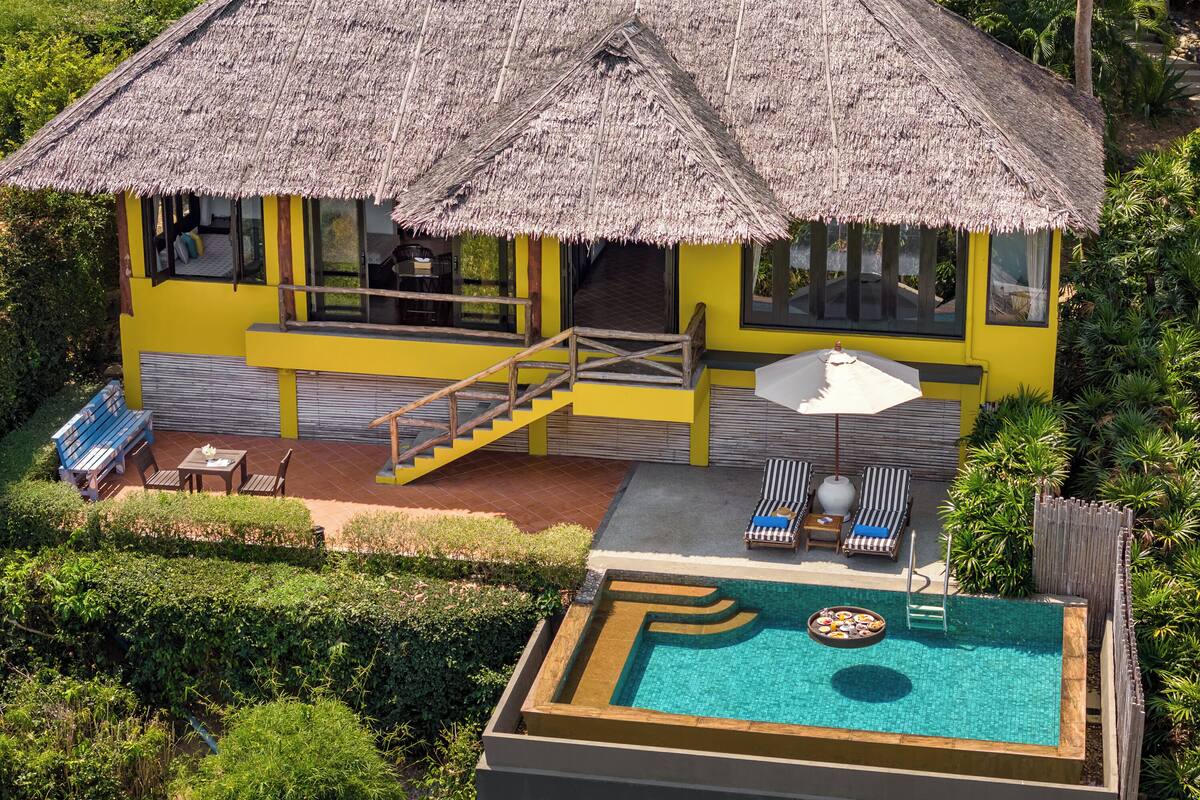 Un bâtiment jaune avec un toit de chaume, une piscine et un escalier menant à un balcon.