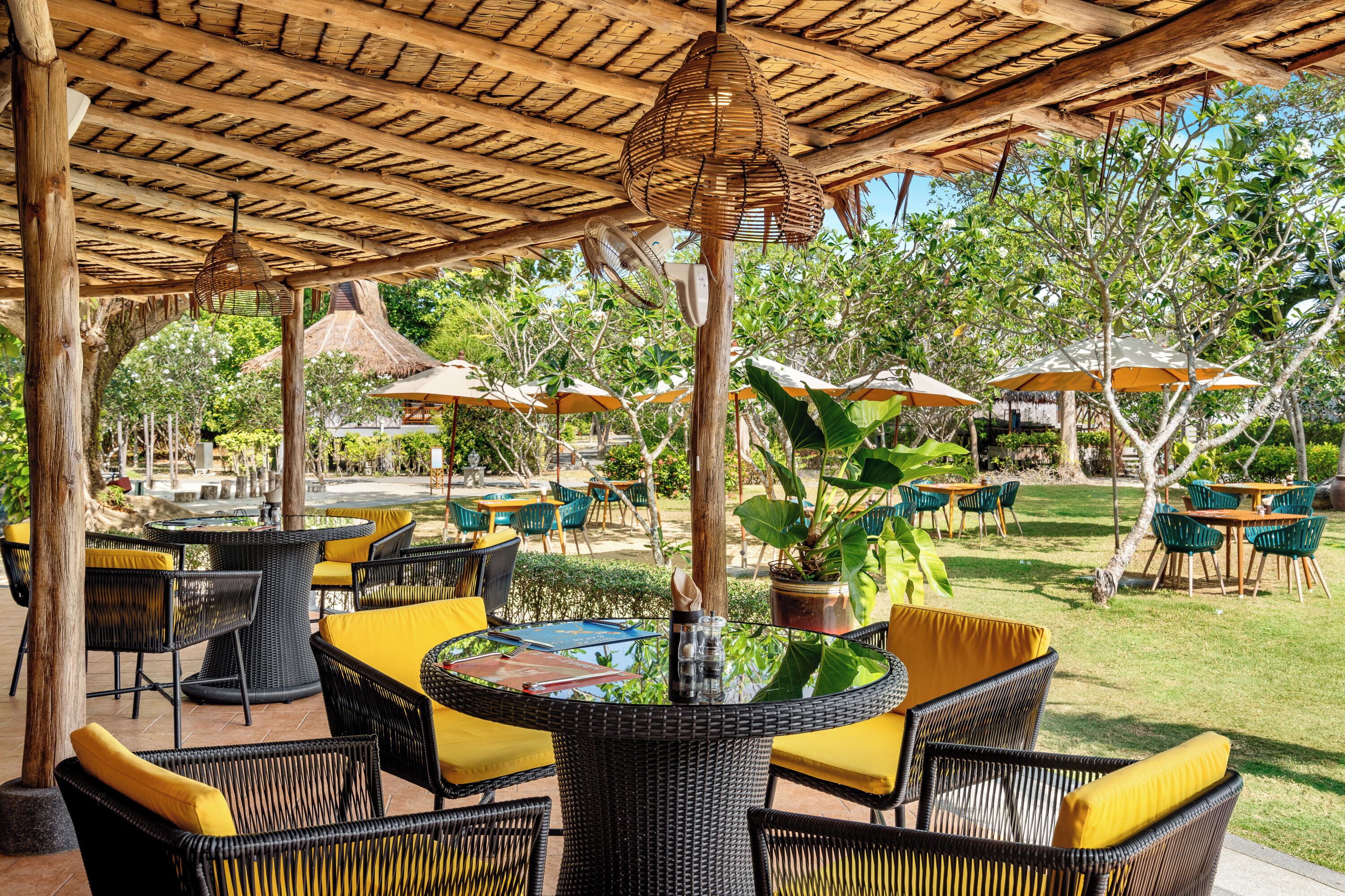 An outdoor patio with wicker furniture and yellow cushions, surrounded by trees and umbrellas.