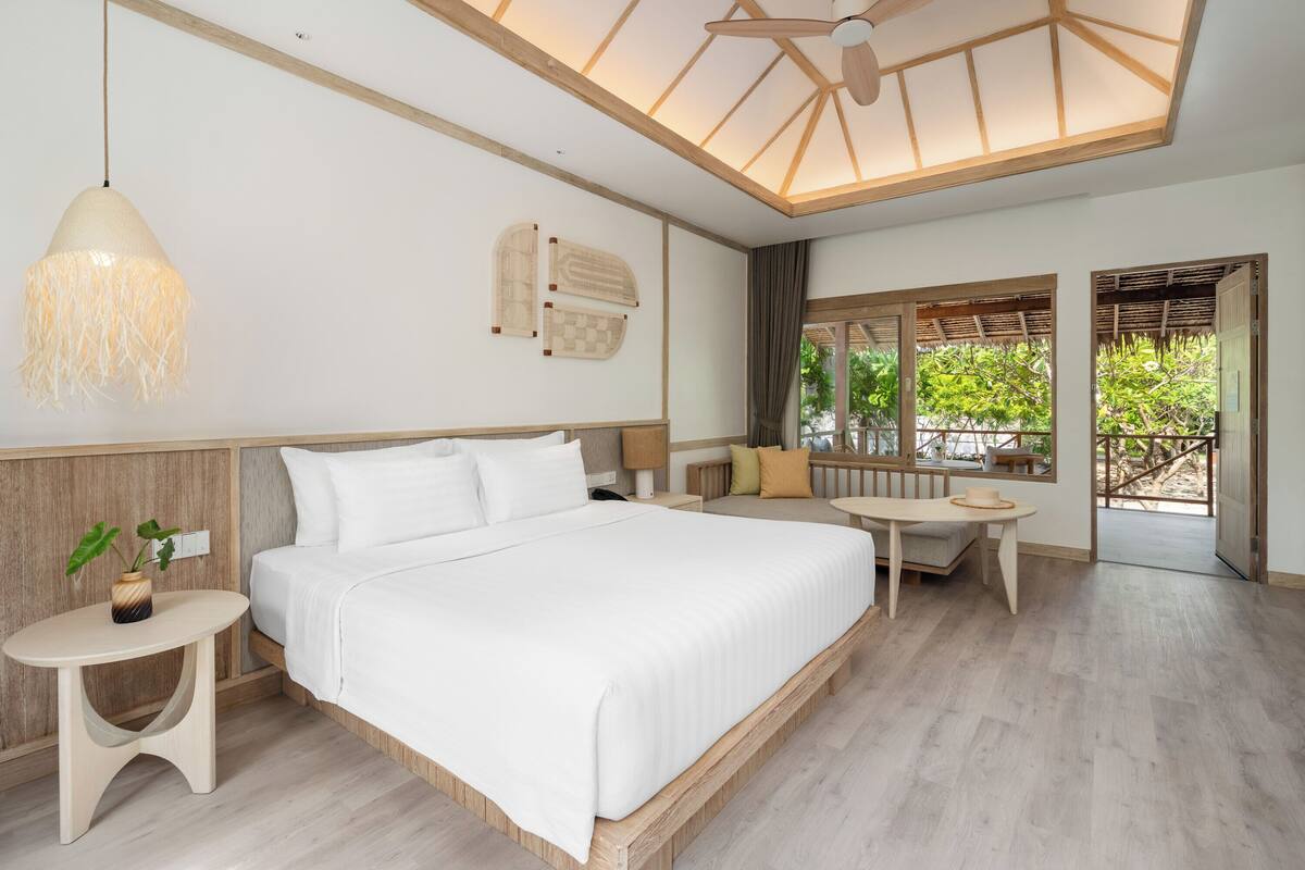 Une chambre spacieuse avec un grand lit, une table de chevet agrémentée d’une plante, un plafond en bois et une vue sur l’extérieur par une fenêtre.
