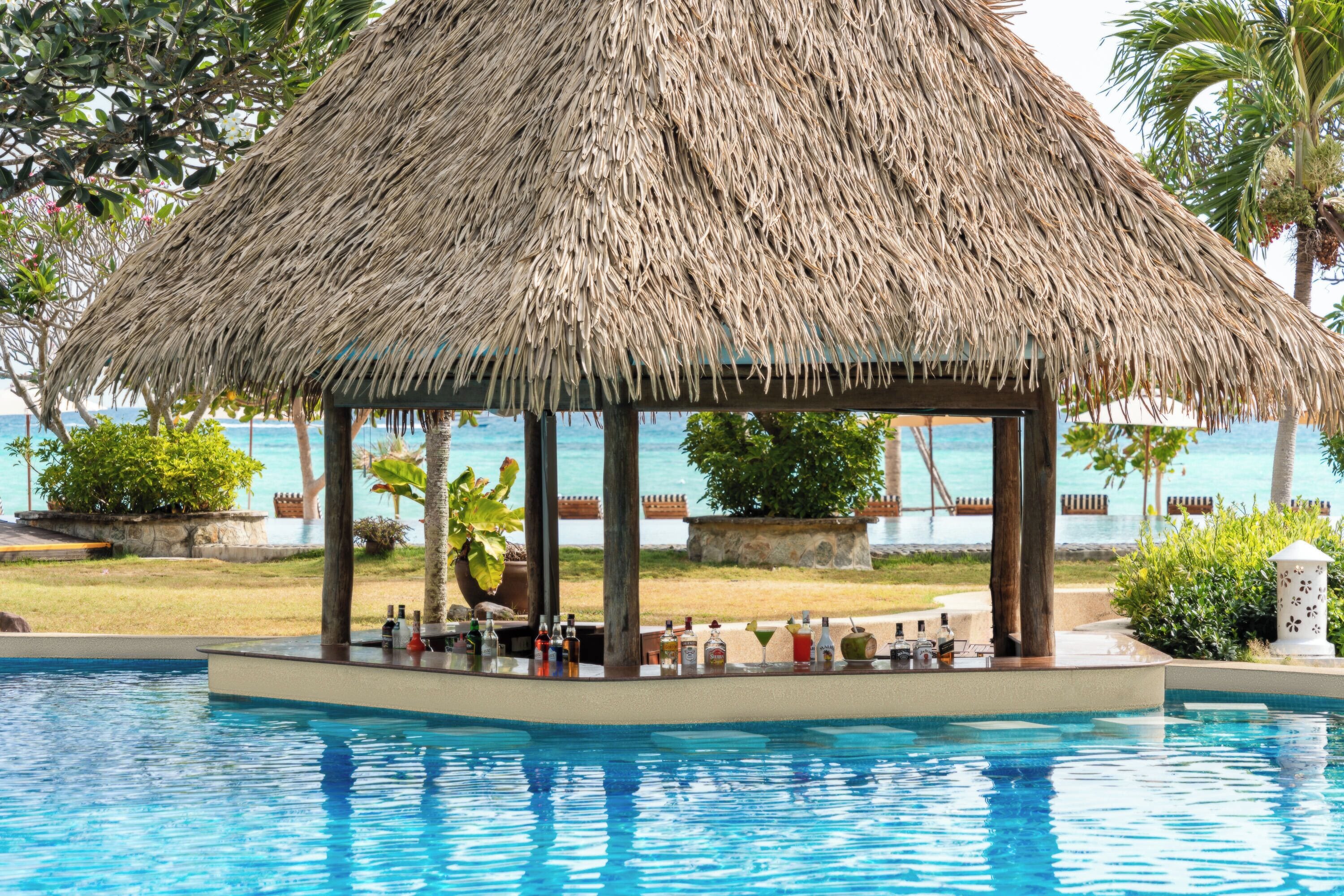 A poolside bar area with a thatched roof structure, surrounded by a pool and palm trees.