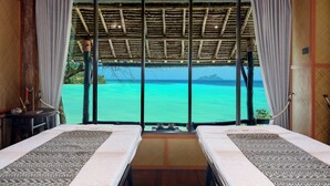 Una habitación con dos camas, vista a aguas turquesas y techo de paja.